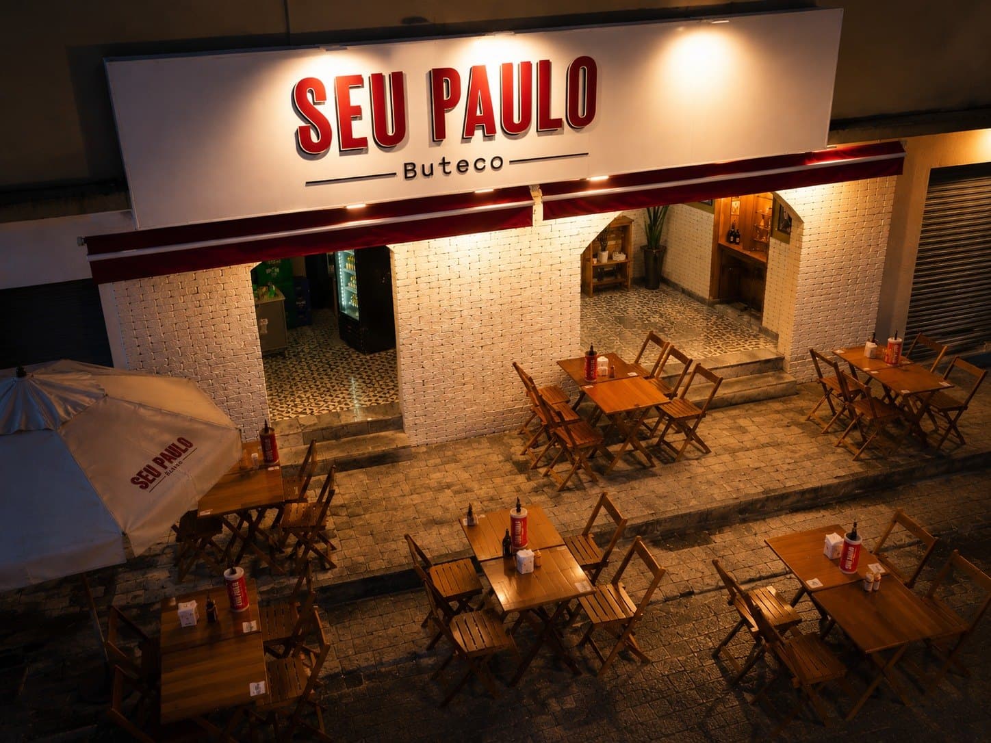 Fachada iluminada do Seu Paulo Buteco à noite, com as mesas na calçada